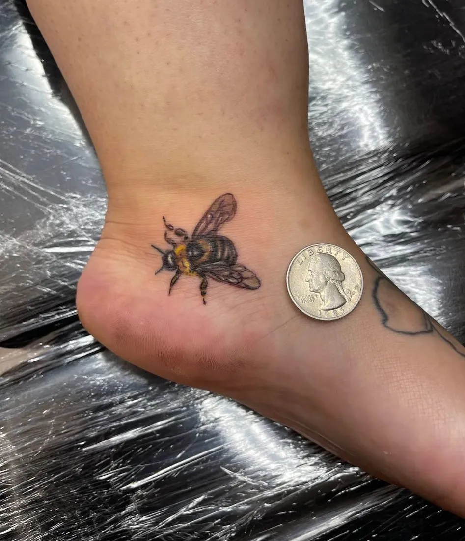 Small realistic bee tattoo on the foot next to a quarter for scale